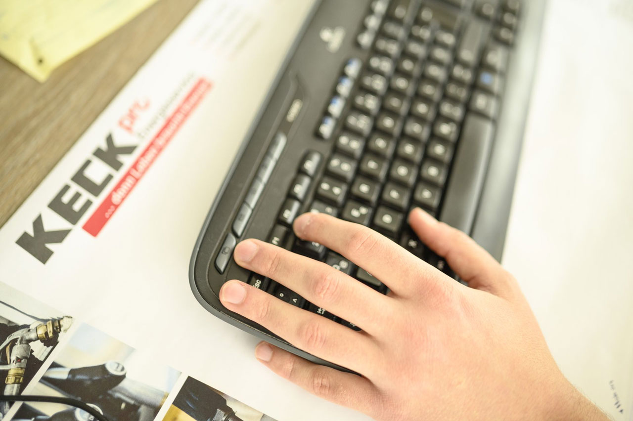 Eine Nahaufnahme einer Hand, die auf eine Computertastatur tippt. Im Hintergrund ist ein unscharfer Ausdruck mit dem Wort 'KECK' erkennbar.