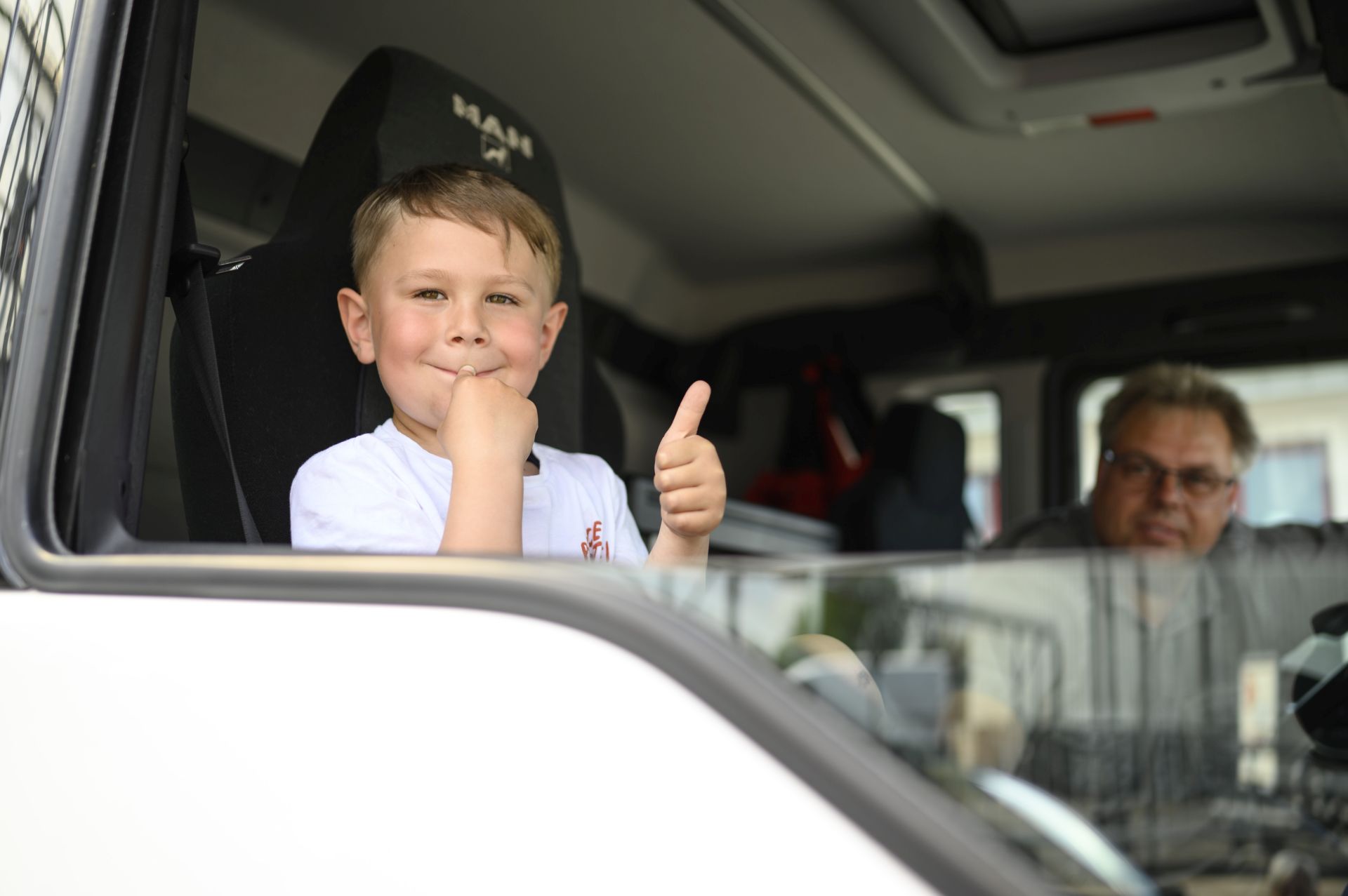 Ein lächelnder Junge mit einem weißen T-Shirt sitzt auf dem Fahrersitz eines Lastwagens und gibt mit seiner rechten Hand einen Daumen hoch. Im Hintergrund ist ein unscharfer Mann zu sehen, der mit im LKW sitzt.