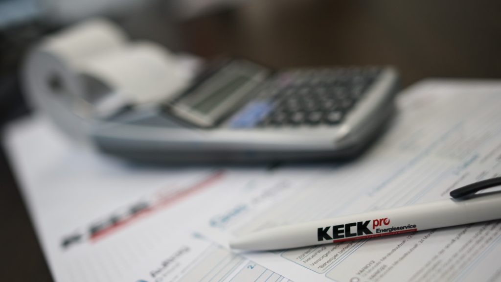 Weißer Kugelschreiber mit Aufdruck 'KECK pro Energieservice' liegt auf Formularen, im unscharfen Hintergrund sind ein Taschenrechner und eine Bonrolle zu erkennen.