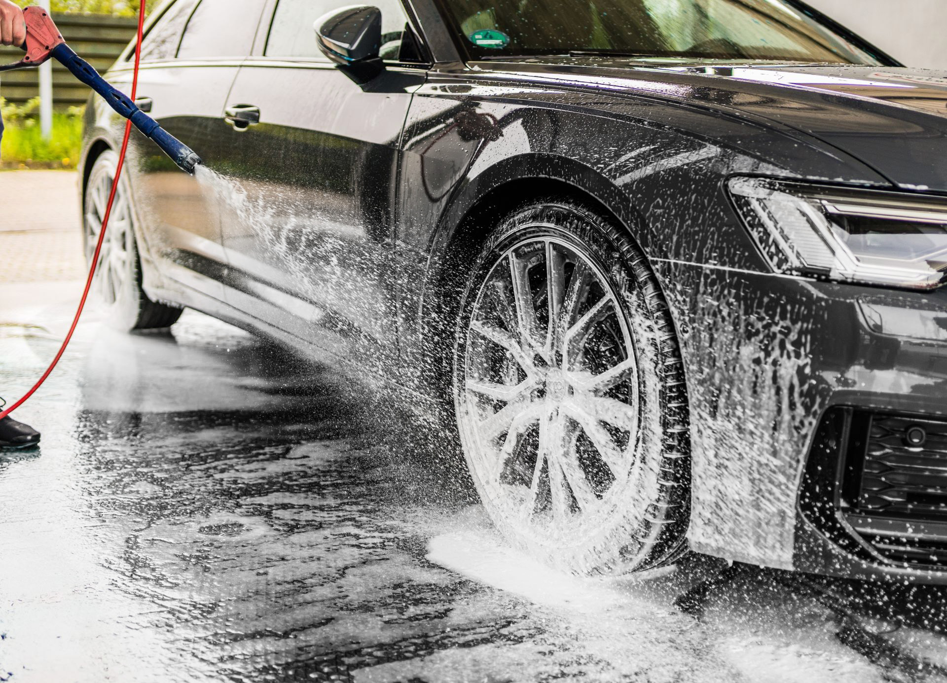 Nahaufnahme eines schwarzen, modernen Autos, an dem eine Person mit einem Hochdruckreiniger Schaum und Wasser über das vordere Rad und die Seitenverkleidung spritzt.